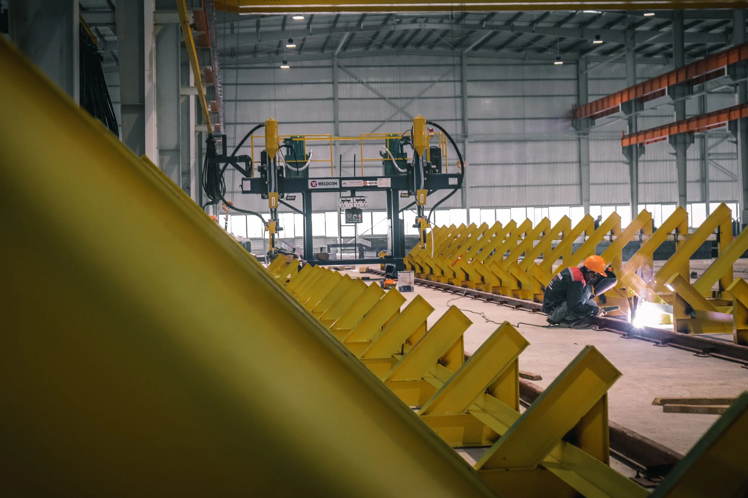 Ouvrier soudant des poutrelles métalliques jaunes alignées dans un hall d'usine.